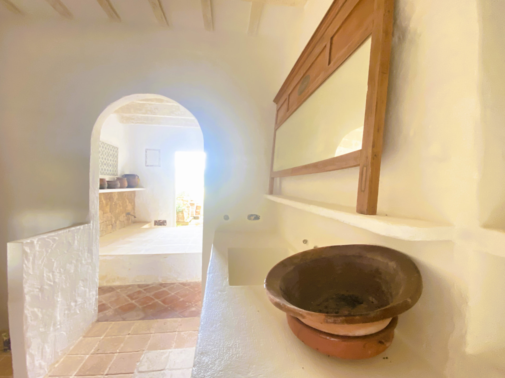 Interior de una finca rústica de Menorca: encimera de obra encalada con pila de barro, espejo de madera y arco de paso a otra estancia con baldosas de barro cocido
